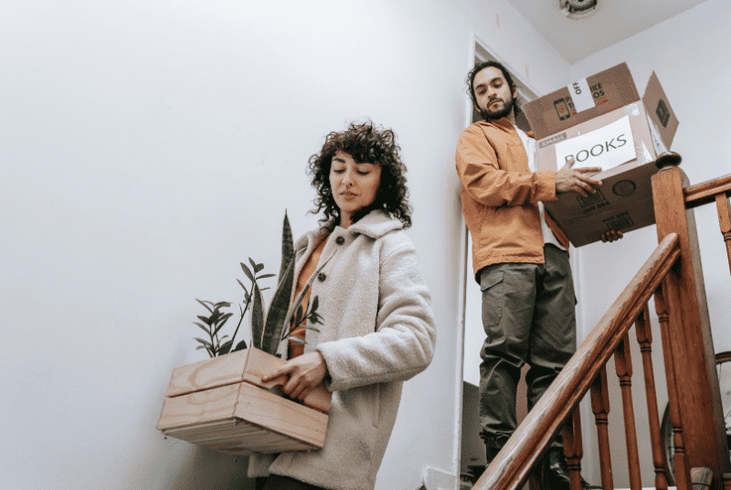 a woman going down the stairs while holding a box of plants and a guy holding a box of books.